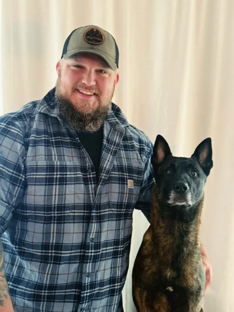 Jacob Robinson in a plaid shirt and baseball cap smiles next to a dark brindle Dutch Shepherd or Belgian Malinois K-9