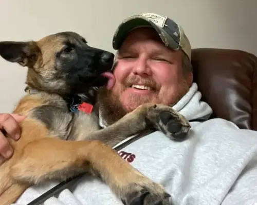 Jacob Robinson lying down smiles as a young Belgian Malinois or German Shepherd puppy licks his face.